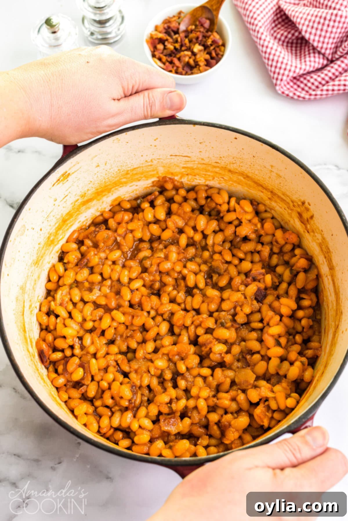 These homemade baked beans are baked in a delicious tangy sweet and savory sauce of brown sugar, maple syrup, ketchup, and apple cider vinegar. A Dutch oven filled with homemade baked beans, being carried by oven-mitted hands to a table, ready to be enjoyed.