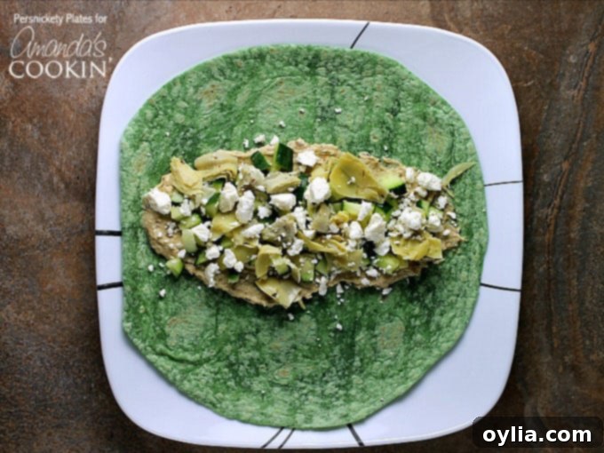 An overhead photo of chopped artichoke hearts, diced cucumber, hummus and crumbled feta artfully arranged on a spinach wrap, ready to be folded. This image showcases the fresh, colorful ingredients.