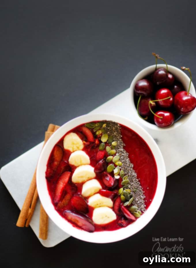 An overhead photo of a stone fruit smoothie in a white bowl topped with sliced plums, bananas cherries, mixed nuts and seeds, showcasing a beautiful, vibrant breakfast.