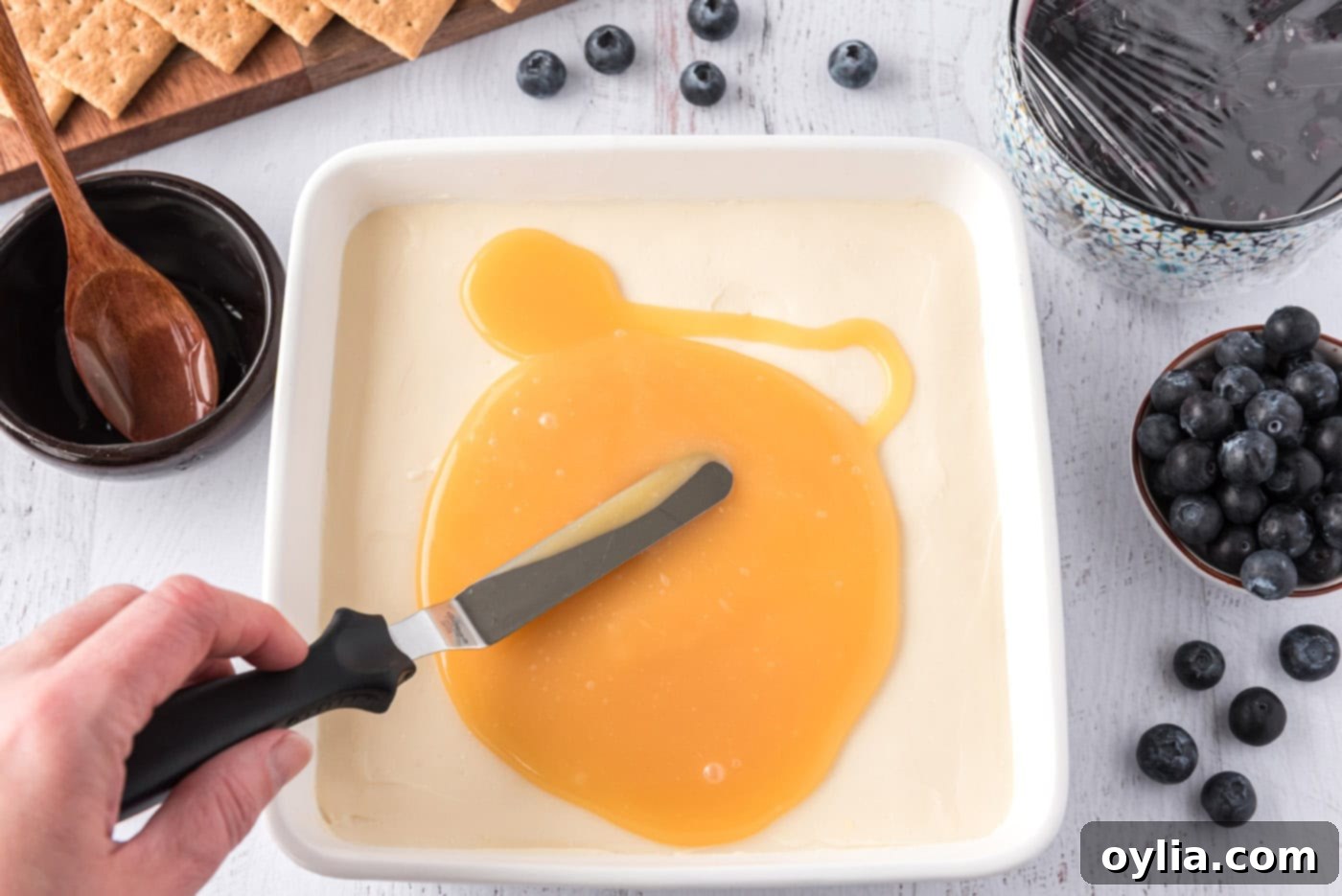 Spreading vibrant lemon curd over the perfectly chilled mascarpone cheesecake layer in a square pan