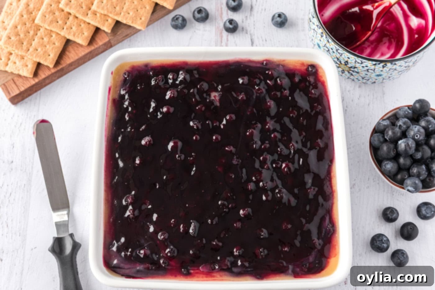 Lush blueberry pie filling spread over the lemon curd and cheesecake layers in a square pan, ready to chill