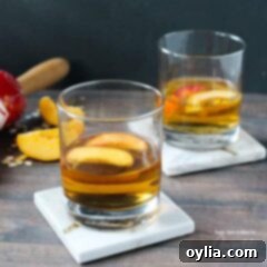 A close-up of two short clear glasses, each brimming with a golden peach pie cocktail, garnished with two fresh peach slices floating on top, ready for enjoyment.