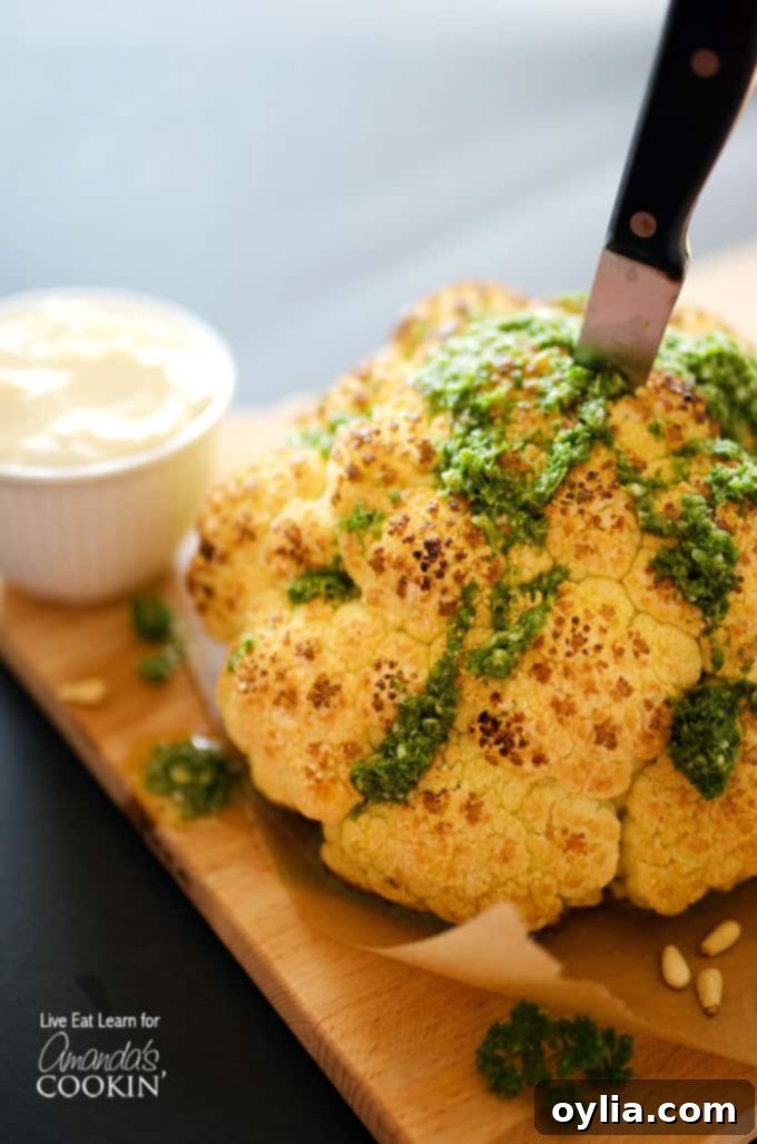 A close up of a whole roasted cauliflower topped with whipped feta and herb sauce on a wooden cutting board with a knife in the center.