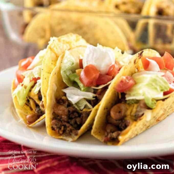 Three hard shell beef and bean oven tacos resting on a white plate topped with lettuce, tomatoes and sour cream.