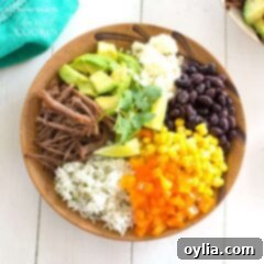 An overhead of a brown bowl filled with white rice, bell pepper, corn, black beans, crumbled queso fresco, avocado, lime and cooked beef.