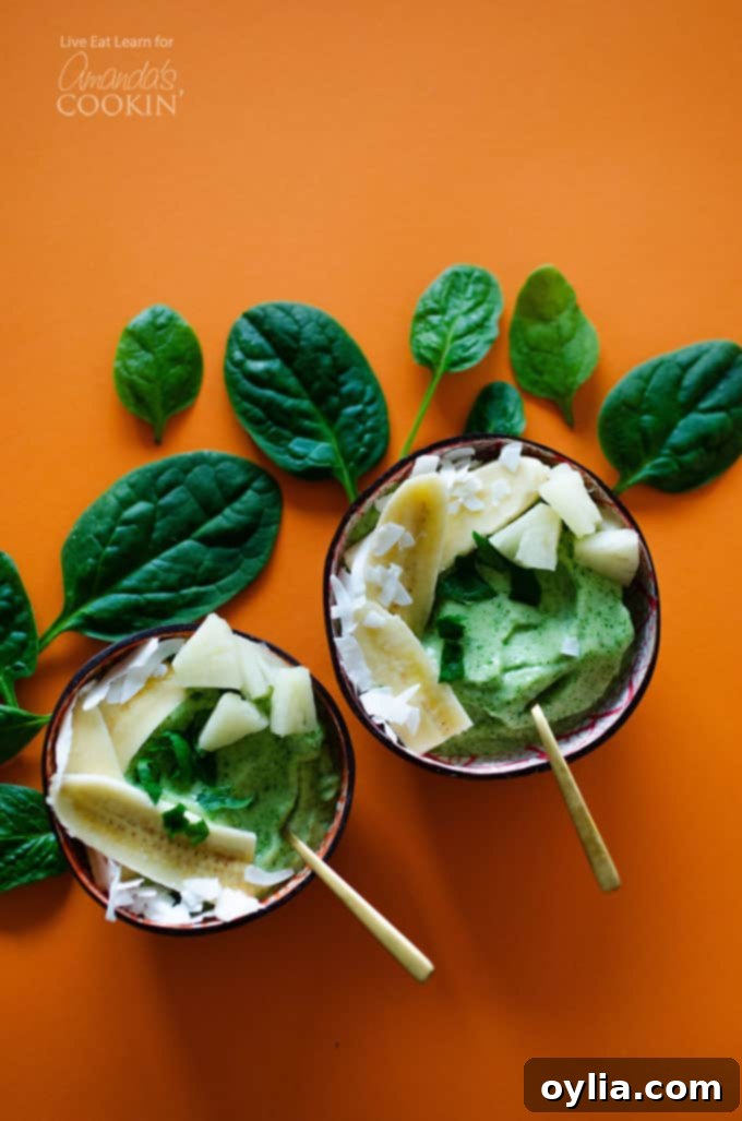 An overhead view of two vibrant green pineapple spinach smoothie bowls, garnished with fresh spinach leaves and other toppings, resting on a rustic wooden table.