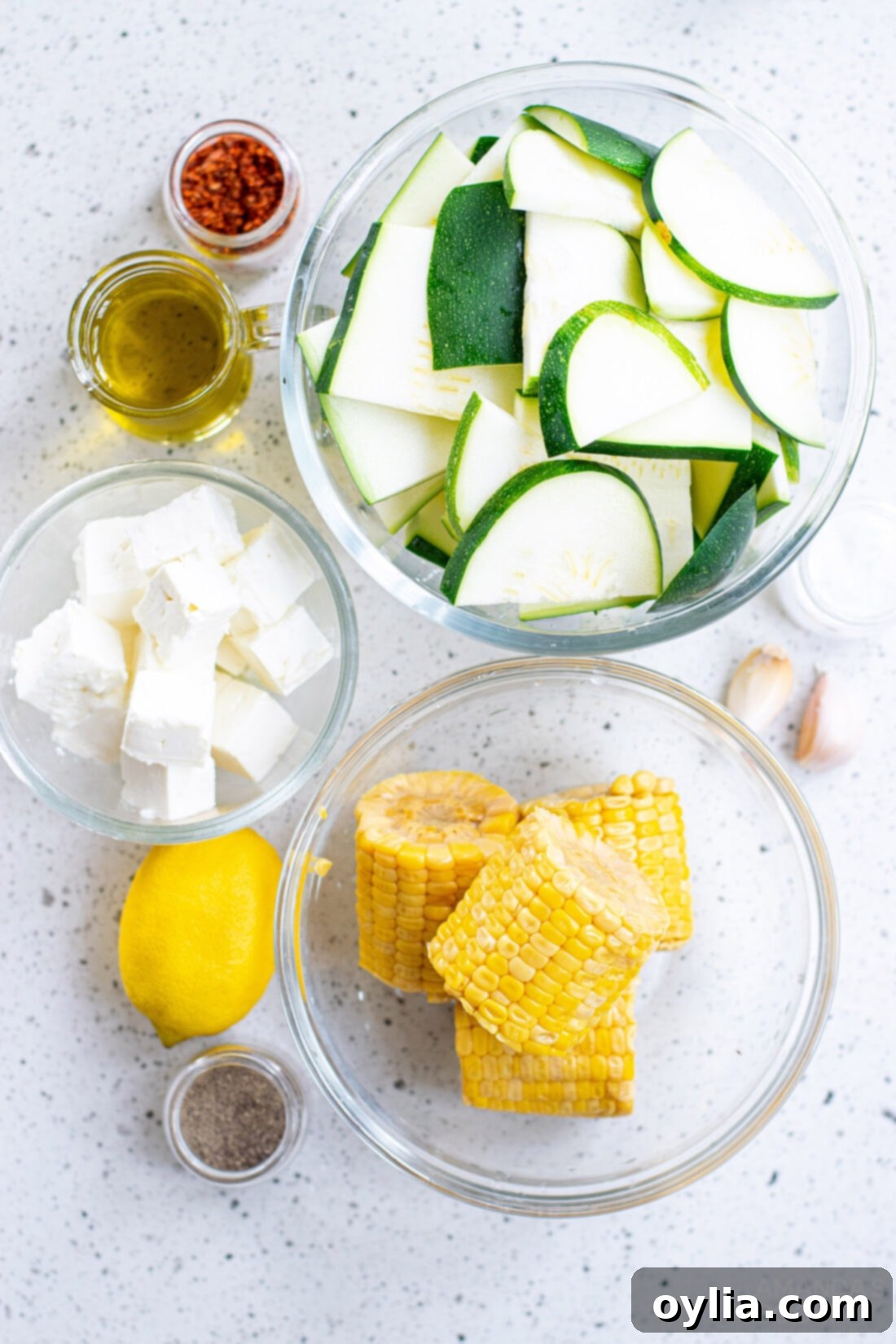 A collection of fresh ingredients laid out for Grilled Corn & Zucchini Salad with Feta, including corn, zucchini, feta, lemon, and spices