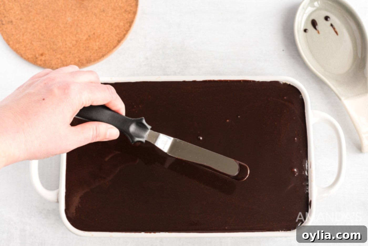 Spreading the warm chocolate ganache smoothly over the top of the eclair cake with an icing spatula, creating a perfect finish.
