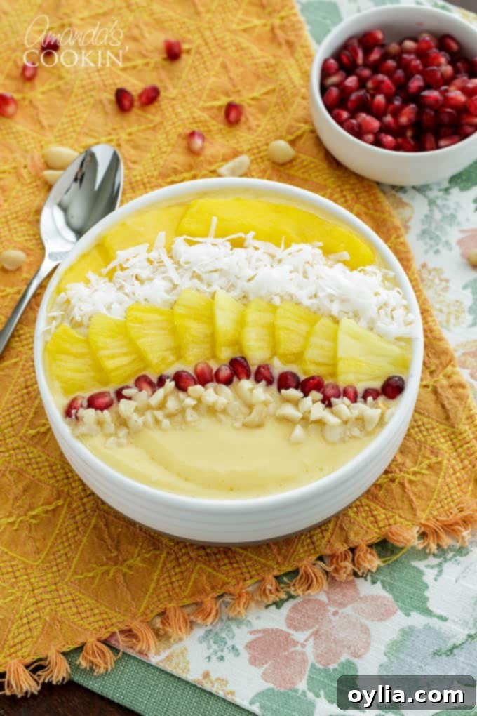 A refreshing tropical smoothie bowl, garnished with fresh mango, pineapple, shredded coconut, and pomegranate arils, sitting on an orange cloth.