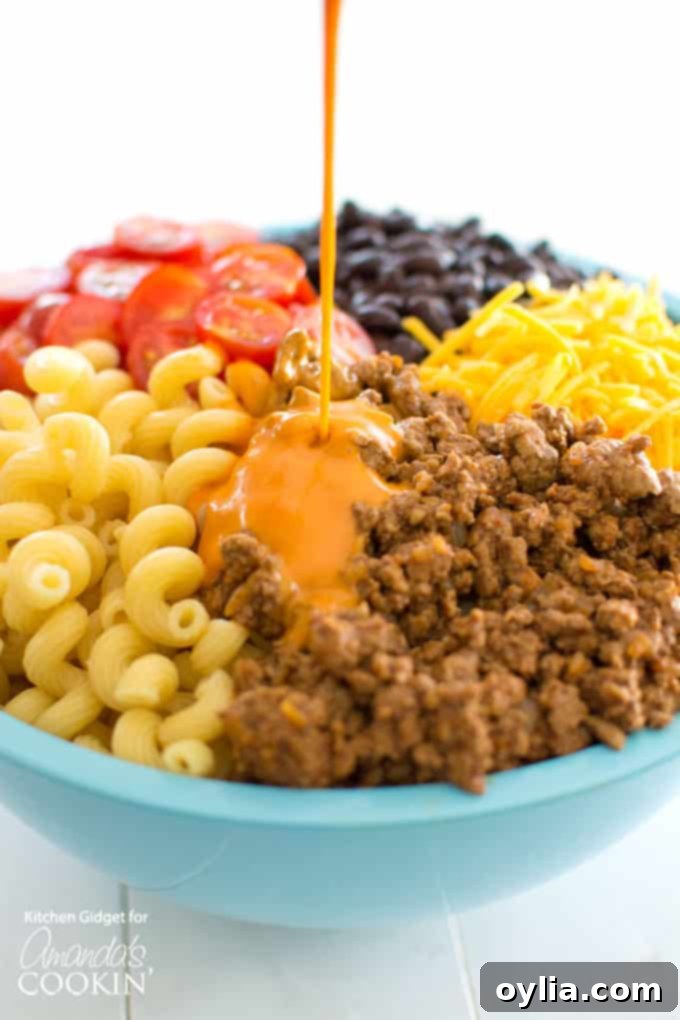 A hand pouring creamy French dressing over the combined ingredients of taco pasta salad in a large mixing bowl.