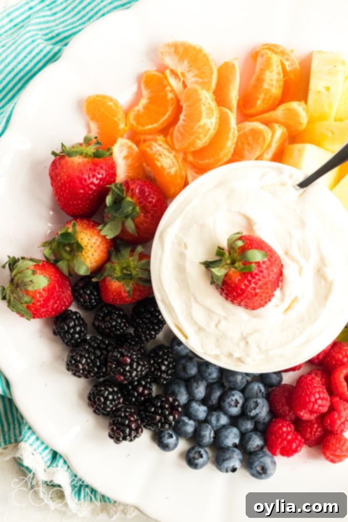 A white platter generously filled with an assortment of colorful fresh fruits like strawberries, blueberries, grapes, and sliced apples, with a bowl of fluffy cream cheese fruit dip positioned invitingly in the center.