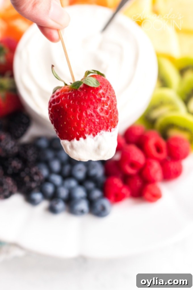 A hand holding a vibrant red strawberry on a toothpick, poised to dip into a creamy, white fruit dip in a bowl.