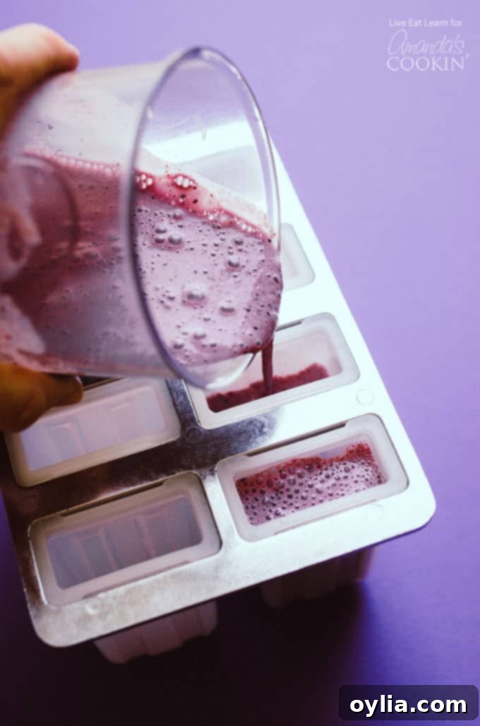 Pouring the creamy blueberry cheesecake popsicle mixture into a silicone popsicle mold, showing the smooth, purple texture before freezing.