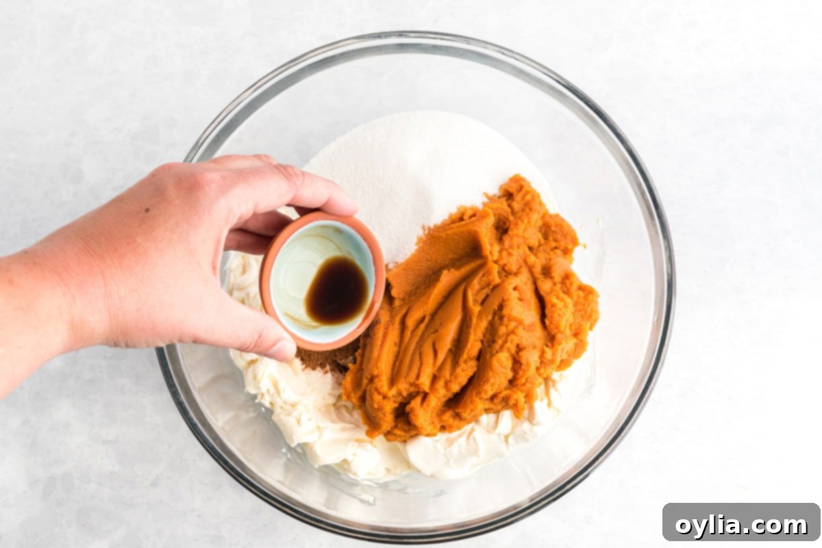 Cream cheese, pumpkin puree, sugar, and spices being added to a mixing bowl to create the cheesecake filling.