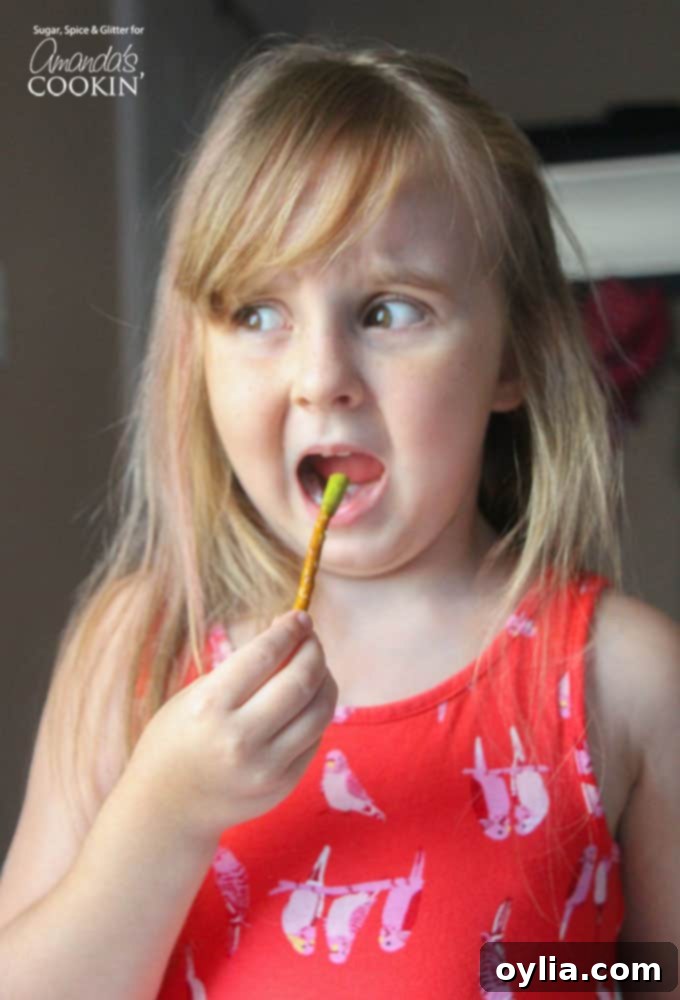 A little girl holding and eating a 'Booger on a Stick' pretzel, dipped in green-tinted Velveeta cheese, at a Halloween party.