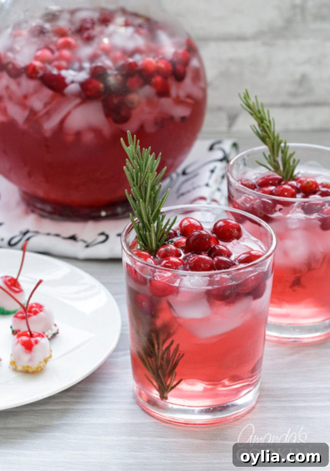 Two glasses and a pitcher of vibrant Cranberry Holiday Punch, garnished with rosemary and cranberries, perfect for festive gatherings.