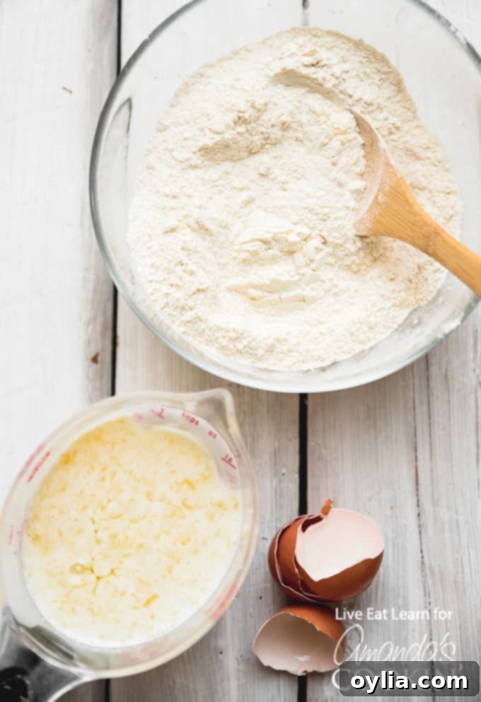 A rustic wooden table displaying all the fresh ingredients needed to make homemade buttermilk pancakes: flour, sugar, baking soda, salt, buttermilk, eggs, and melted butter.