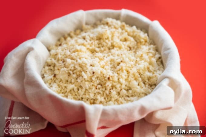 Grated cauliflower wrapped in a cheesecloth, being vigorously squeezed by hands over a bowl to extract all excess moisture.