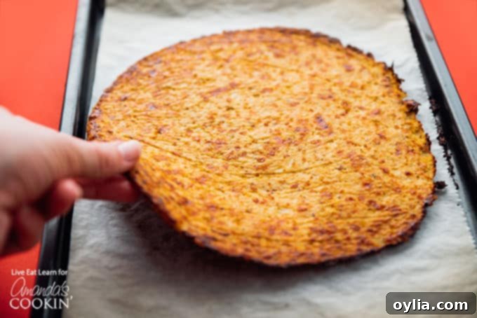 A golden-brown cauliflower pizza crust, pre-baked and ready for toppings, being gently lifted from the parchment paper.