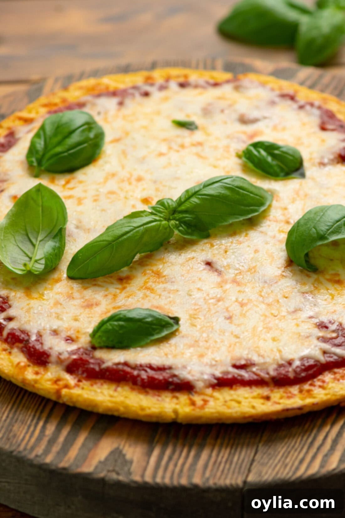 A vibrant cauliflower pizza crust topped with melted cheese, fresh green basil leaves, and a sprinkle of spices, presented on a wooden board.