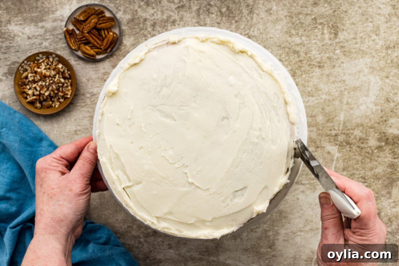 A bench scraper being used to remove excess cream cheese frosting from the sides of a carrot cake on a turntable, creating a crumb coat.