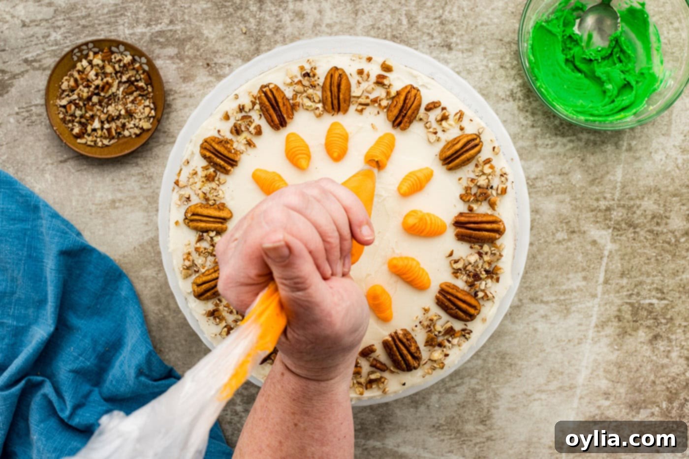 A hand expertly piping orange frosting in the shape of a carrot onto the top of a frosted carrot cake.