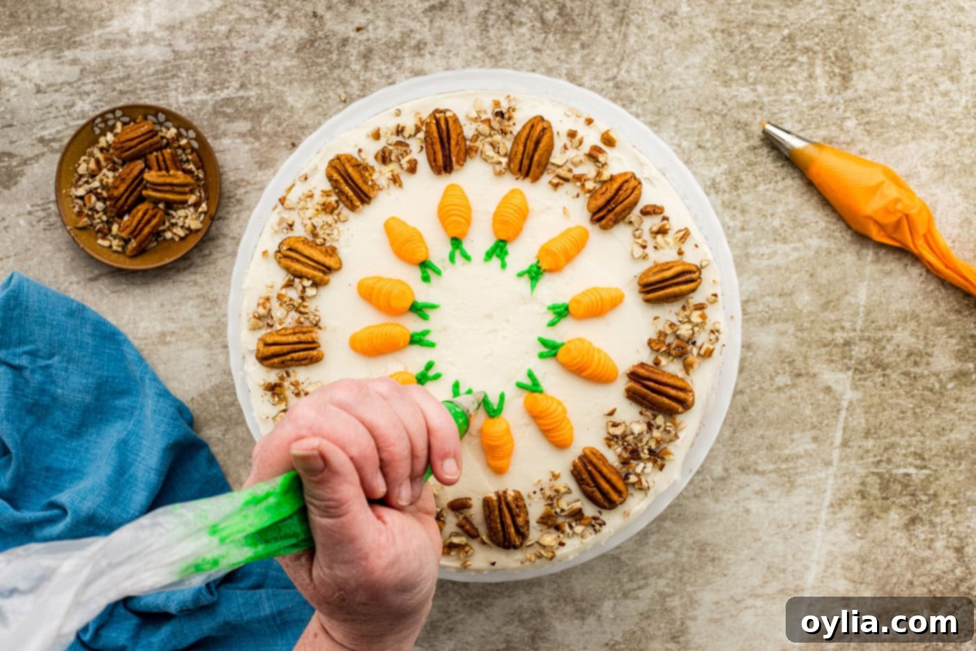 A hand piping small green frosting leaves onto the top of a carrot cake, complementing the orange piped carrots.