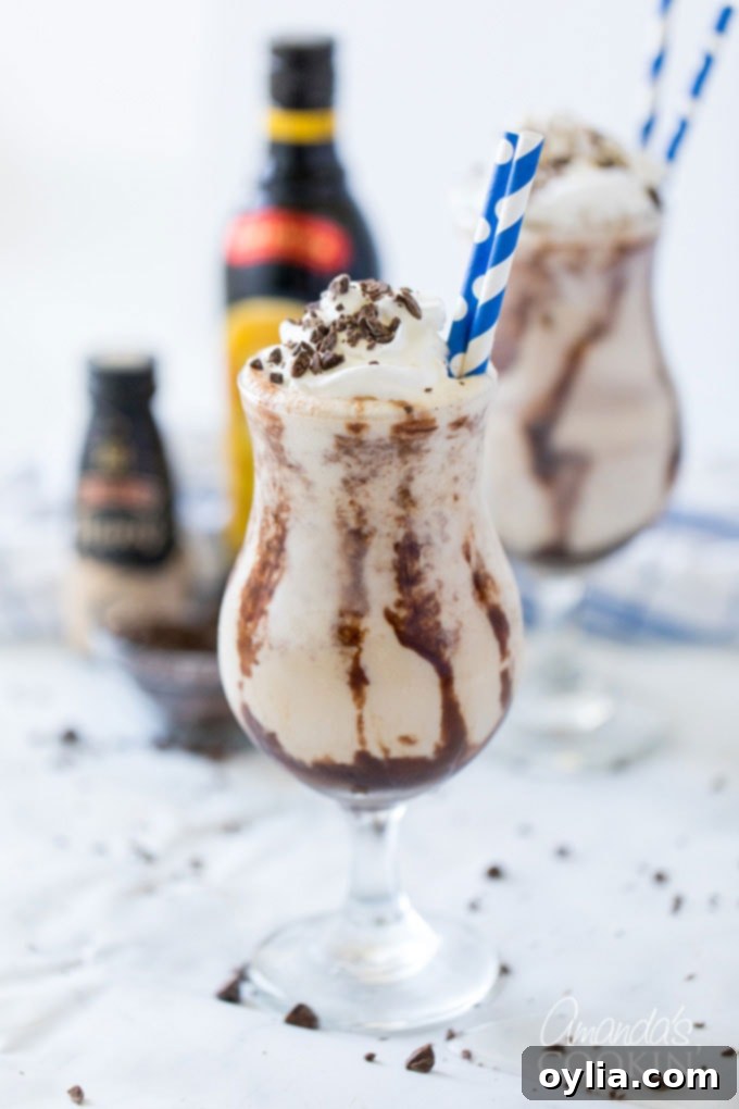 Two perfectly prepared Frozen Mudslide cocktails in hurricane glasses, with bottles of Kahlua, Baileys, and vodka blurred in the background.