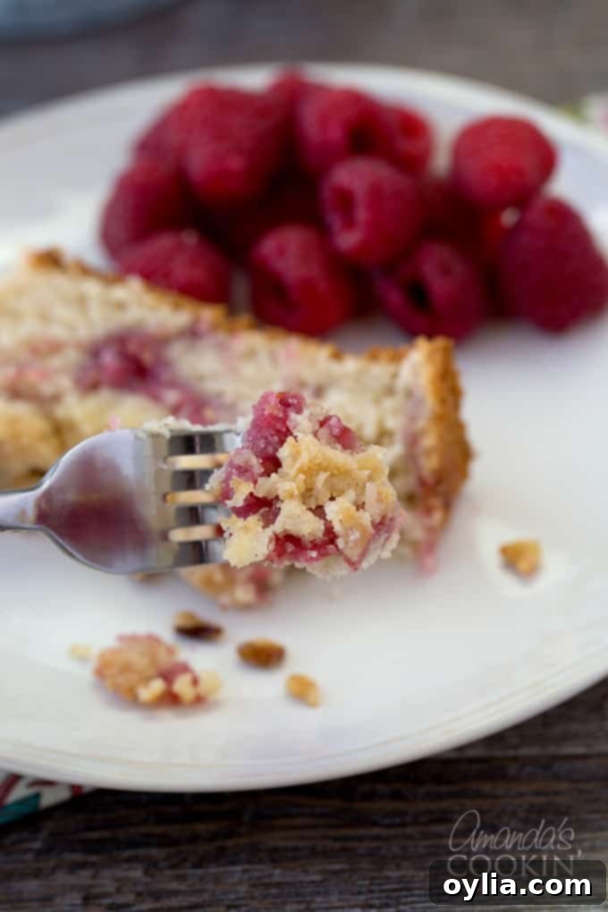 A close-up shot of a fork taking a bite of raspberry coffee cake, showcasing the tender crumb and glistening raspberry filling, with a whole slice in the background.