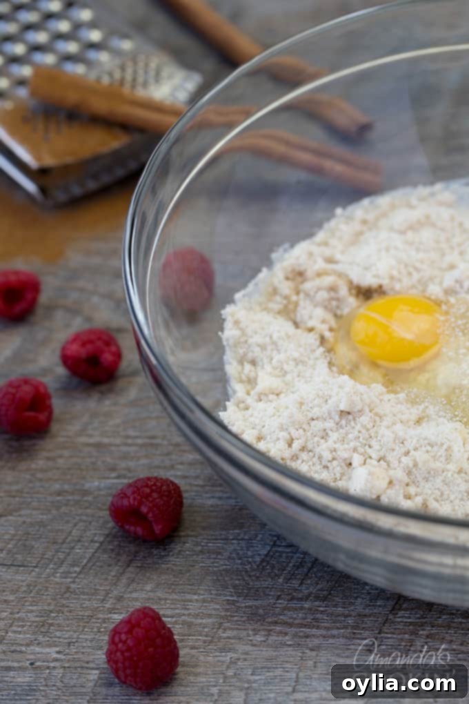 Fresh, vibrant raspberries next to a clear bowl containing the flour mixture and a freshly cracked egg yolk, poised for the next step of the recipe.