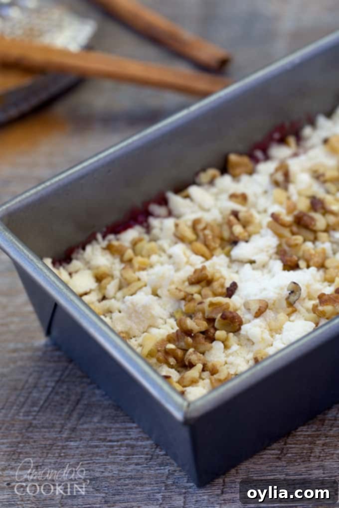 A close-up view of the loaf pan, showcasing layers of tender batter, vibrant raspberry filling, a generous sprinkle of crumb topping, and crunchy chopped nuts.
