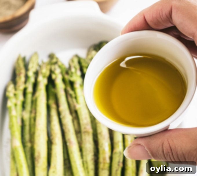 A hand drizzling olive oil from a small dish over fresh asparagus spears in a larger bowl, ready for seasoning.