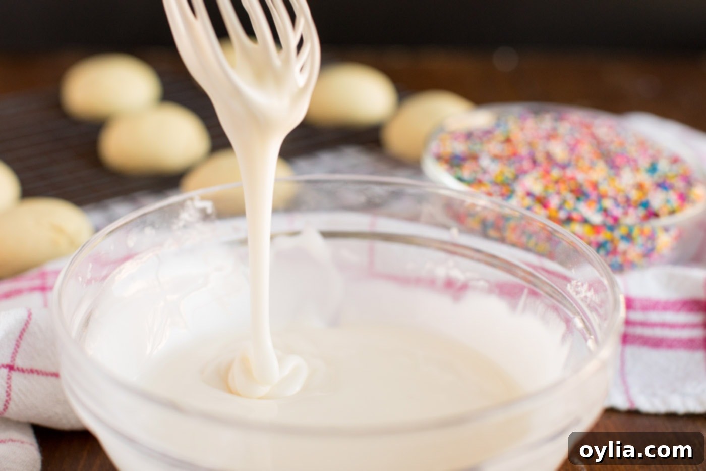 Close-up of a whisk lifting thick, smooth white glaze from a bowl, perfectly demonstrating the ideal consistency for coating Italian Anisette Cookies.