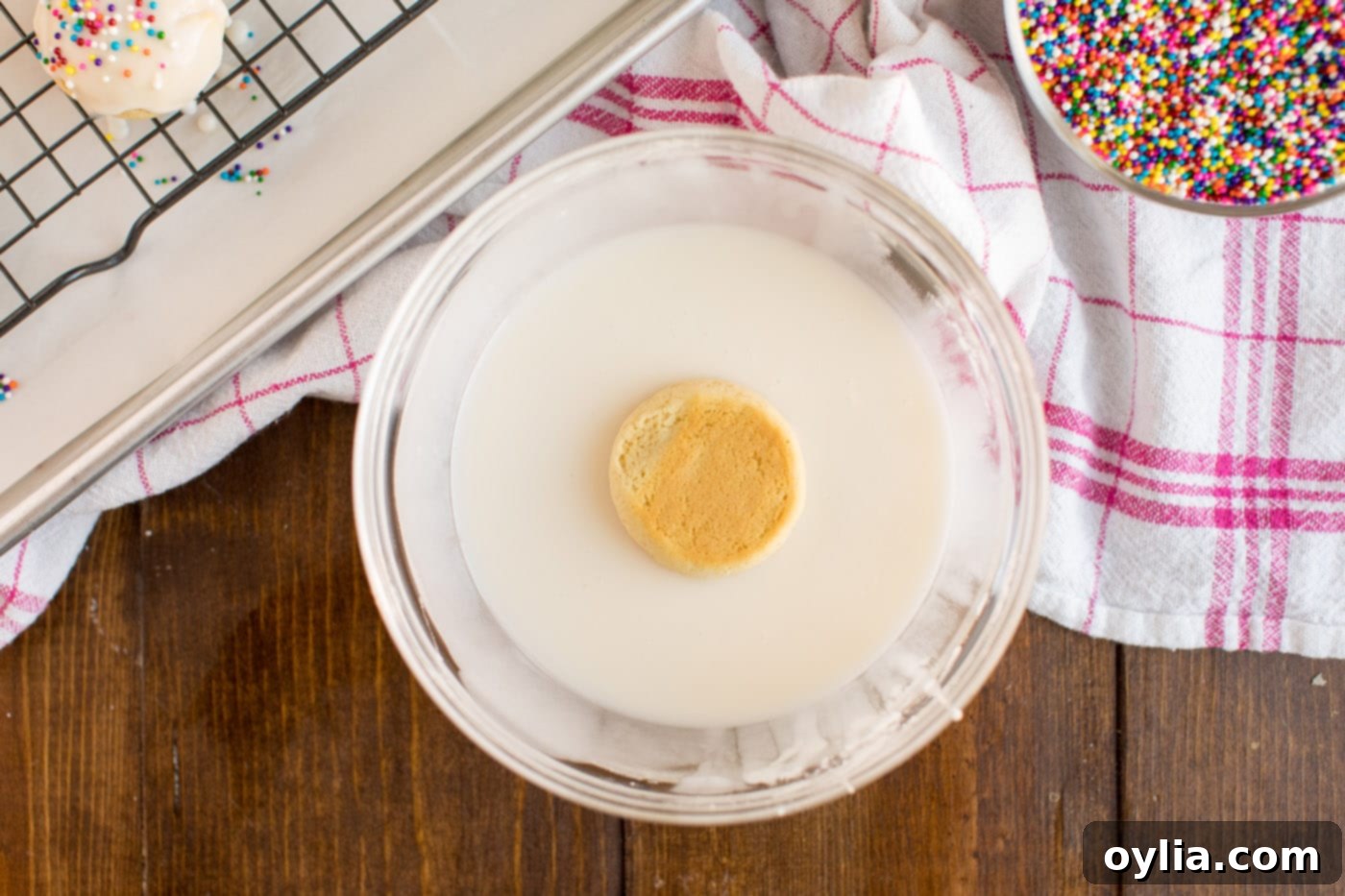 A hand gently dipping a freshly baked Italian Anisette Cookie into a bowl of thick white glaze, preparing it for its colorful sprinkle topping.