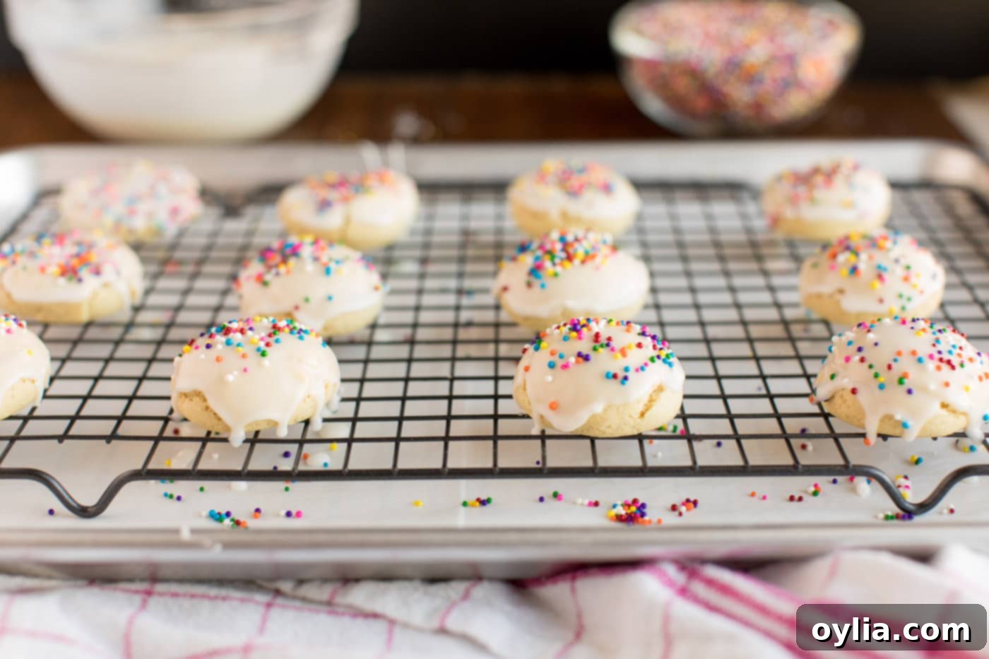 A group of freshly glazed Italian Anisette Cookies, glistening with their white glaze and topped with a scattering of colorful sprinkles, neatly arranged on a wire rack.
