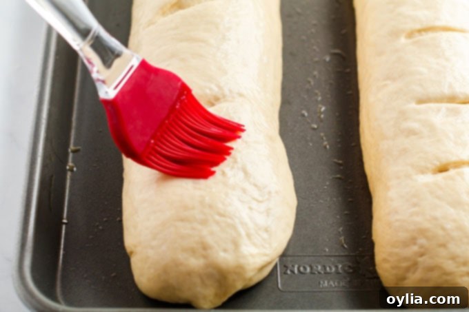 Close-up of a pastry brush applying egg wash to the top of a risen Italian bread loaf before baking.