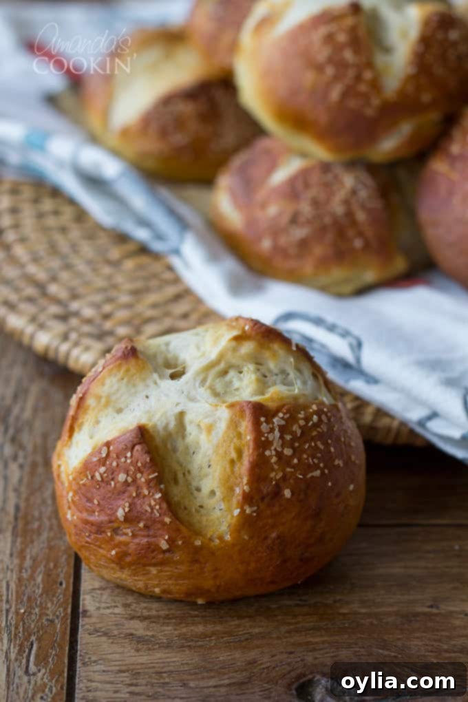 A close up of a soft homemade bretzel roll with other golden brown pretzel buns laying on a white and blue towel in the background.