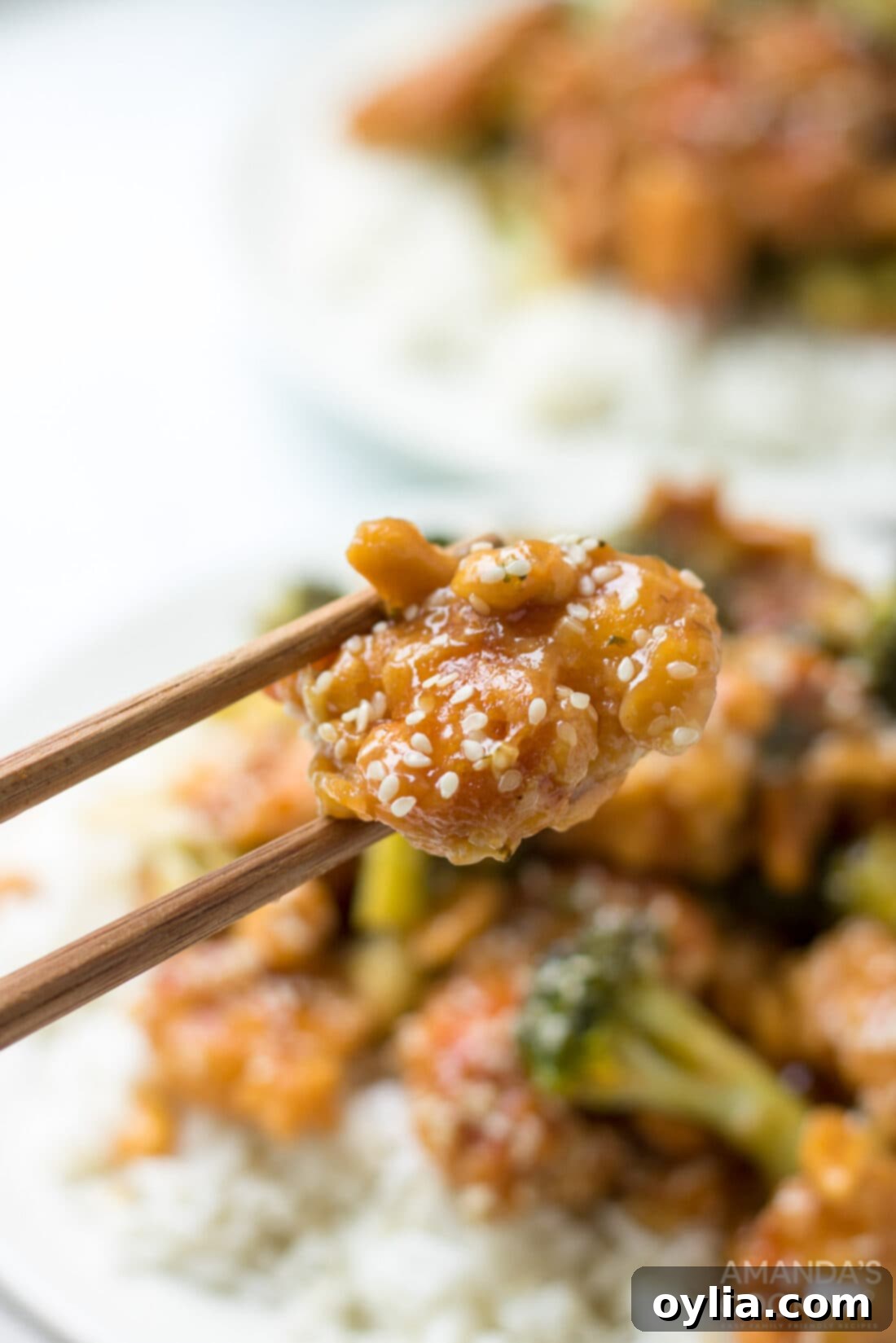 Sesame chicken is complemented by a mixture of diced ginger, red wine vinegar, hoisin sauce, soy sauce, and topped with broccoli and sesame seeds. chopstick holding sesame chicken piece