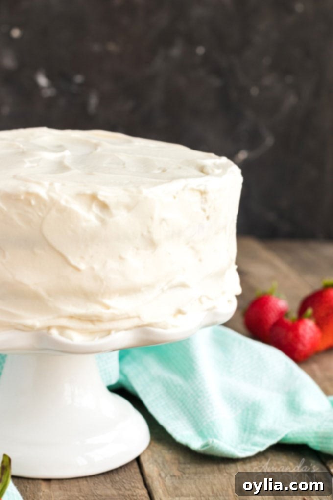 Only 8 ingredients needed for this fluffy white cake recipe that bakes up in just 30 minutes! Perfect for a party, in layers, or baked as a sheet cake! frosted white cake on a cake stand with a blue towel and strawberries in the background