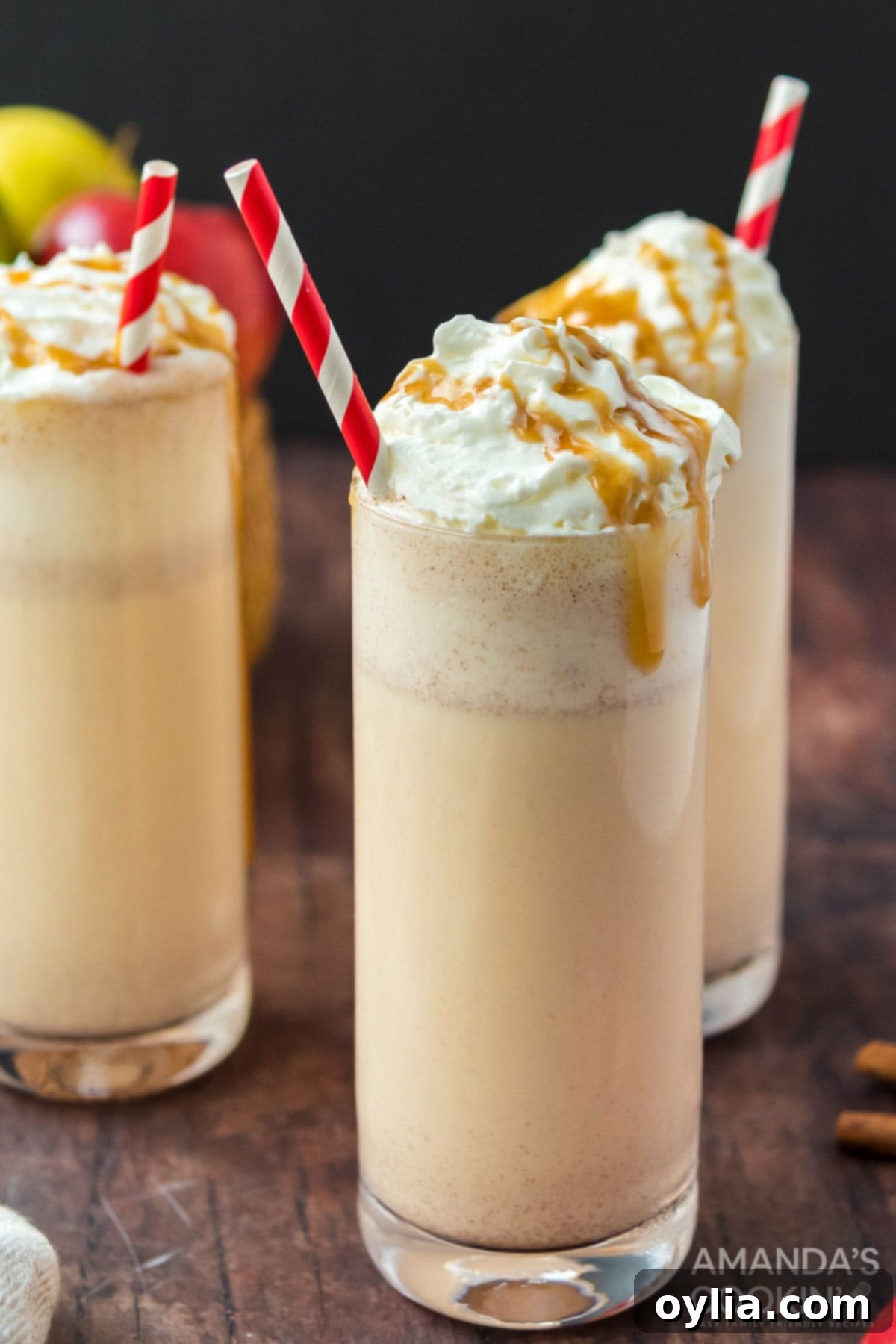 Three glasses of creamy apple cider milkshakes, garnished with whipped cream and a cinnamon stick, against a fall backdrop.