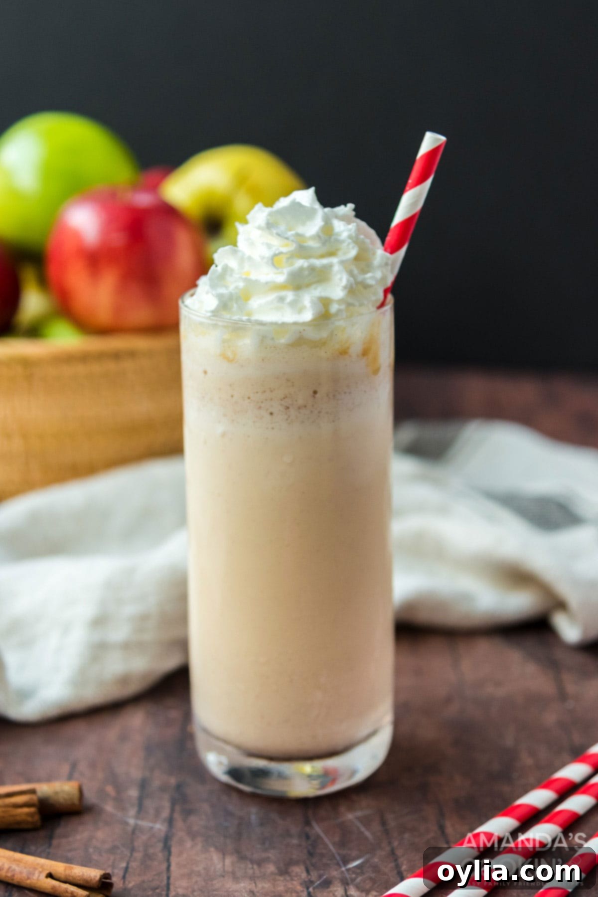 A tall glass of frothy apple cider milkshake with a festive straw, topped with whipped cream and a sprinkle of cinnamon, against a blurred rustic background.