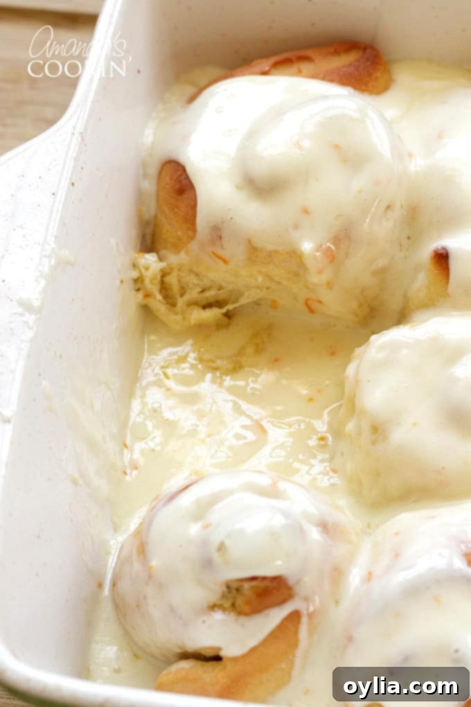 A baking pan of warm, unfrosted orange rolls with one roll already removed, ready for a generous dollop of cream cheese frosting.