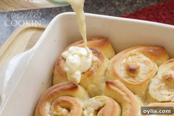 A close-up shot of a creamy, dreamy cream cheese frosting being generously drizzled over a pan of warm orange rolls, fresh from the oven.