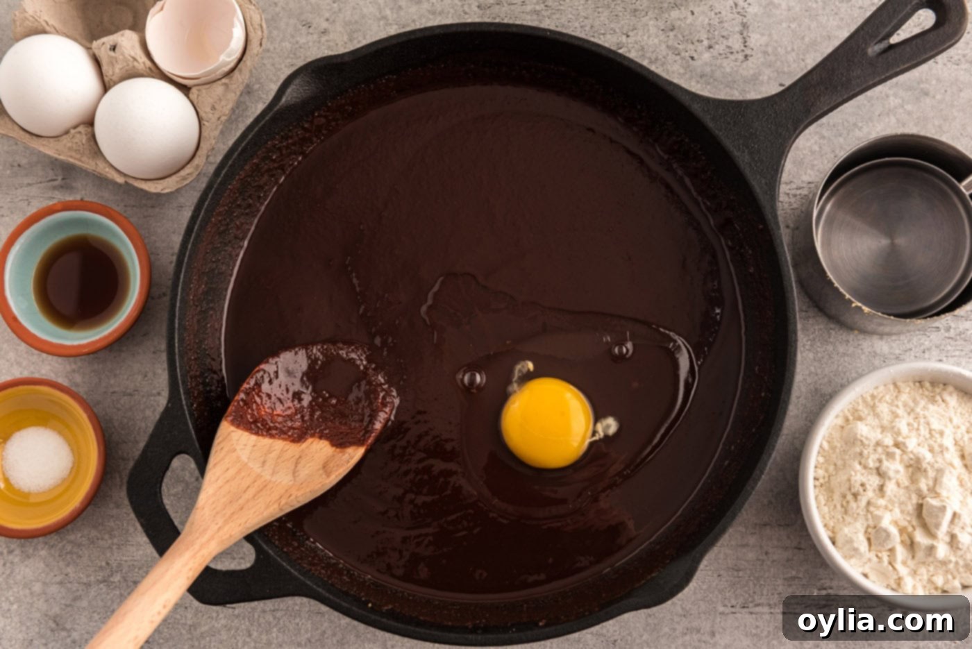 A raw egg being mixed thoroughly into the rich chocolate brownie batter in a cast iron skillet using a wooden spoon, ensuring full incorporation.