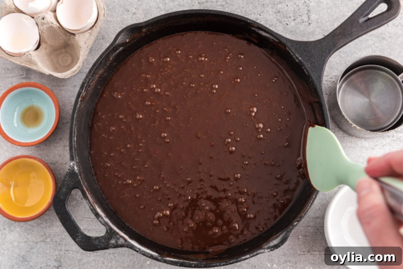 A rubber spatula meticulously scraping down the edges of the cast iron skillet, gathering all the rich brownie batter into the center for even baking.