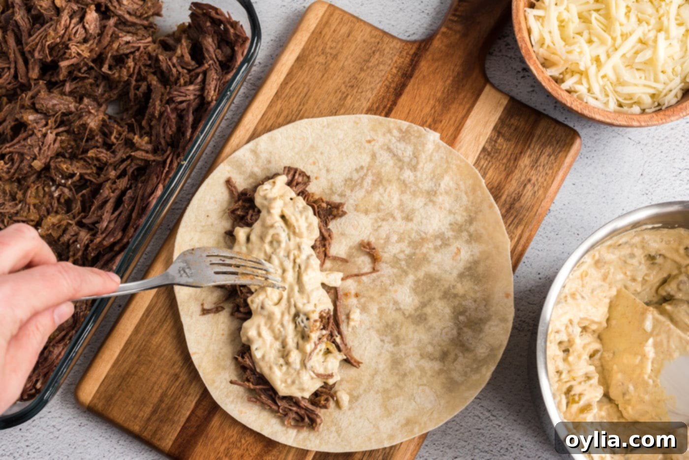 A generous scoop of the creamy green chile sour cream mixture being added over the shredded beef filling on a flour tortilla.