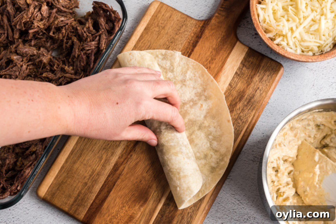 Carefully rolling up a flour tortilla filled with shredded beef and green chile sour cream mixture, preparing it for baking.