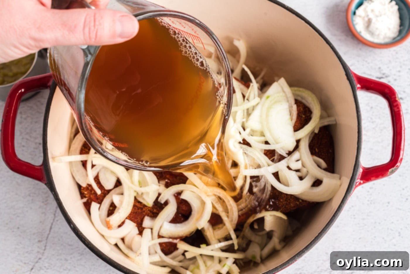 Pouring rich beef broth and white wine vinegar over the chuck roast and onions in a Dutch oven, preparing for roasting.