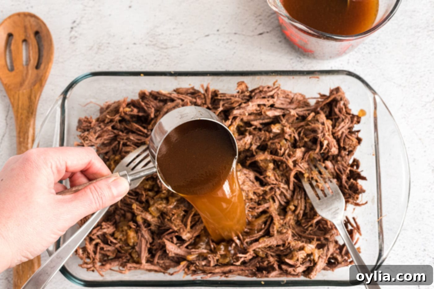 Pouring rich beef pan juices over the shredded beef and onion mixture to moisten the filling, enhancing its flavor and texture.