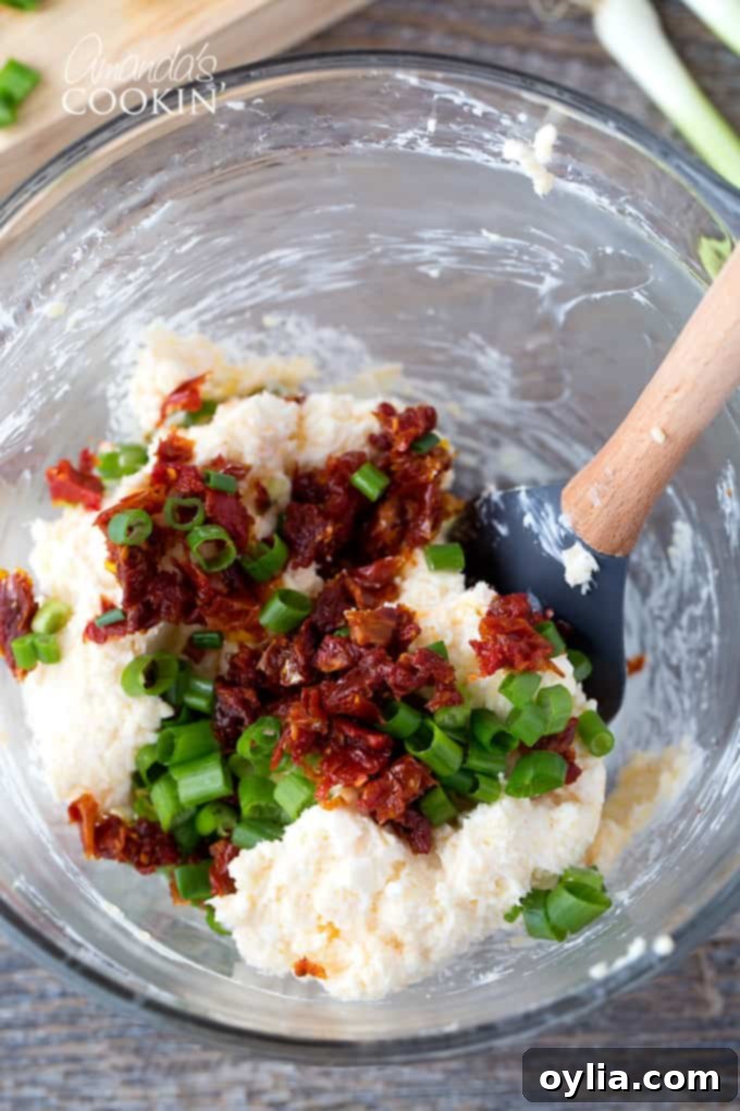 A bowl of creamy cheese mixture with chopped chives and sun-dried tomatoes visibly mixed in, awaiting the next step.